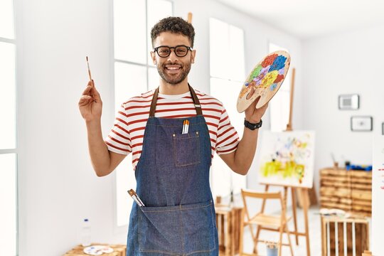 Young arab artist man smiling happy holding paintbrush and palette at art studio.