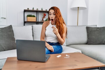 Young redhead woman doing coronavirus infection nasal test scared and amazed with open mouth for...