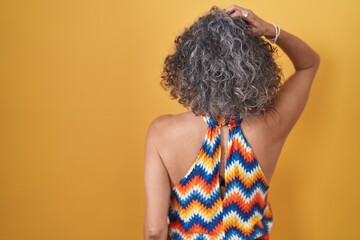 Middle age woman with grey hair standing over yellow background backwards thinking about doubt with hand on head