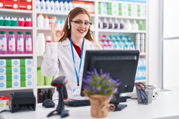 Redhead woman working at pharmacy drugstore wearing headset doing ok sign with fingers, smiling friendly gesturing excellent symbol
