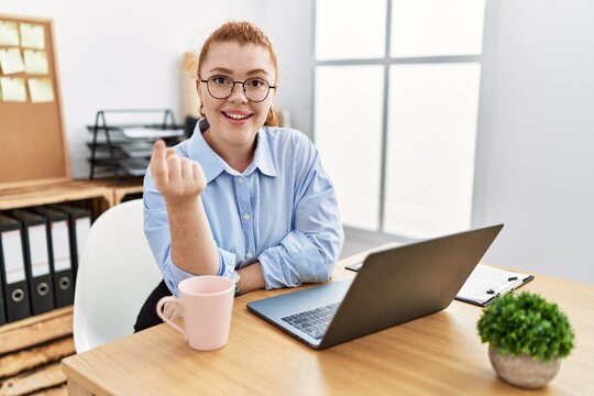 Young redhead woman working at the office using computer laptop beckoning come here gesture with hand inviting welcoming happy and smiling