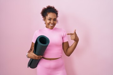 Young hispanic woman with curly hair holding yoga mat over pink background looking confident with...