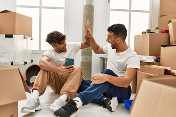 Two hispanic men couple using smartphone high five with hands raised up at new home