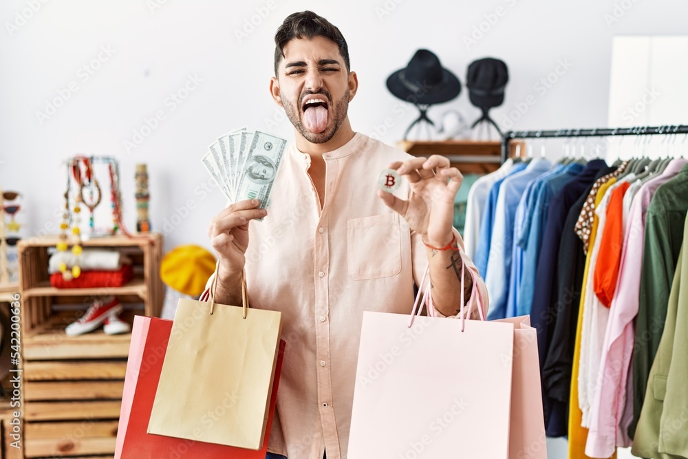 Canvas Prints Young hispanic man holding shopping bags, dollars and bitcoin sticking tongue out happy with funny expression.