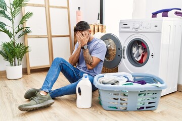 Young hispanic man putting dirty laundry into washing machine with sad expression covering face...
