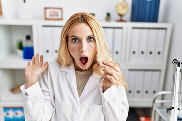 Beautiful blonde dentist woman holding invisible aligner orthodontic at dental clinic scared and...