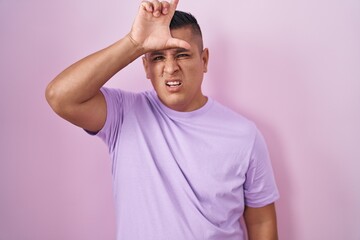 Young hispanic man standing over pink background making fun of people with fingers on forehead...