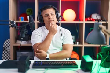 Young hispanic man playing video games with hand on chin thinking about question, pensive expression. smiling and thoughtful face. doubt concept.