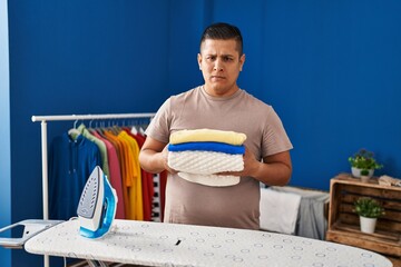 Hispanic young man holding folded laundry after ironing skeptic and nervous, frowning upset because...