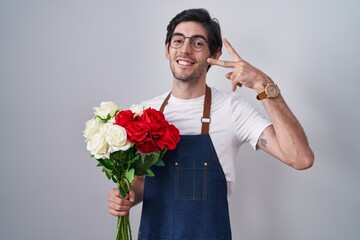 Young hispanic man holding bouquet of white and red roses doing peace symbol with fingers over face, smiling cheerful showing victory