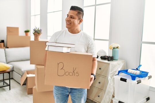 Young latin man smiling happy holding books cardboard box moving at new home.