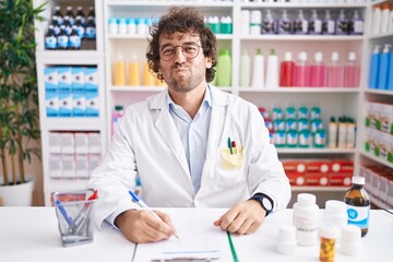 Hispanic young man working at pharmacy drugstore puffing cheeks with funny face. mouth inflated with air, crazy expression.