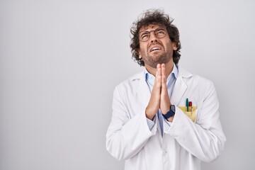 Hispanic young man wearing doctor uniform begging and praying with hands together with hope expression on face very emotional and worried. begging.
