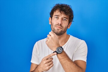 Hispanic young man standing over blue background in hurry pointing to watch time, impatience,...