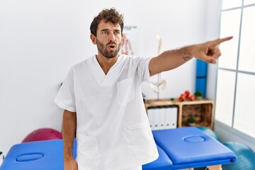 Young handsome physiotherapist man working at pain recovery clinic pointing with finger surprised ahead, open mouth amazed expression, something on the front