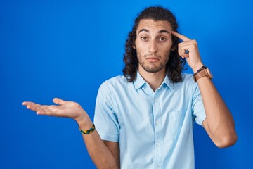 Fototapeta premium Young hispanic man standing over blue background confused and annoyed with open palm showing copy space and pointing finger to forehead. think about it.
