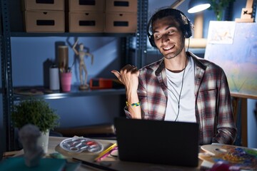 Young hispanic man sitting at art studio with laptop late at night smiling with happy face looking and pointing to the side with thumb up.