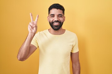 Hispanic man with beard standing over yellow background showing and pointing up with fingers number two while smiling confident and happy.