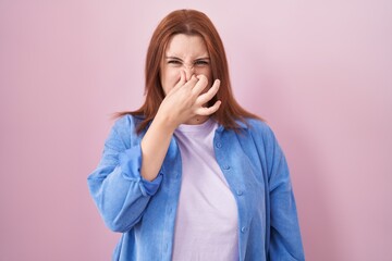 Young hispanic woman with red hair standing over pink background smelling something stinky and...
