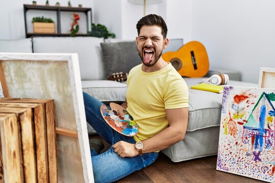 Young man with beard painting canvas at home sticking tongue out happy with funny expression. emotion concept.