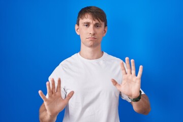 Caucasian blond man standing over blue background moving away hands palms showing refusal and denial with afraid and disgusting expression. stop and forbidden.