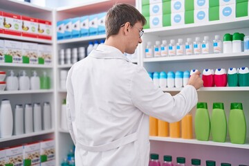 Young caucasian man pharmacist smiling confident holding product of shelving at pharmacy