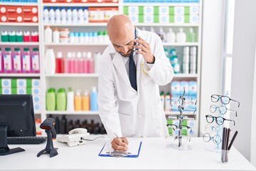 Young bald man pharmacist talking on smartphone writing on document at pharmacy