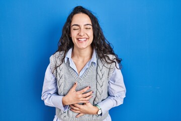Young brunette woman standing over blue background smiling and laughing hard out loud because funny...