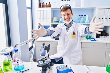 Caucasian man working at scientist laboratory looking at the camera smiling with open arms for hug. cheerful expression embracing happiness.