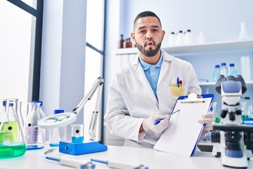 Hispanic man working at scientist laboratory holding blank clipboard making fish face with mouth and squinting eyes, crazy and comical.