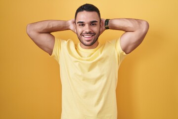 Fototapeta na wymiar Young hispanic man standing over yellow background relaxing and stretching, arms and hands behind head and neck smiling happy