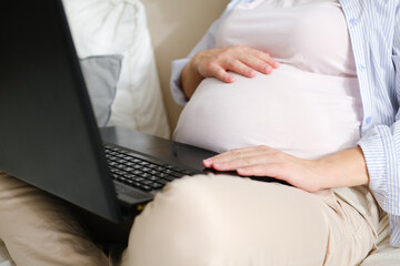 Beautiful young pregnant woman working on a laptop at home sitting on the bed