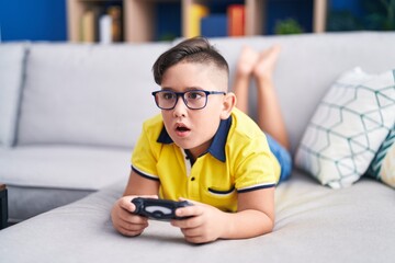Young hispanic kid playing video game holding controller on the sofa in shock face, looking...