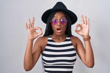 Young african american with braids wearing hat and sunglasses looking surprised and shocked doing ok approval symbol with fingers. crazy expression