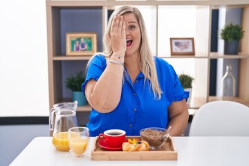 Obraz na płótnie Canvas Caucasian plus size woman eating breakfast at home covering one eye with hand, confident smile on face and surprise emotion.