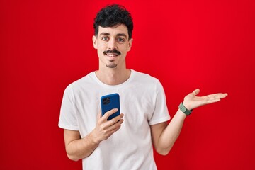 Hispanic man using smartphone over red background pointing aside with hands open palms showing copy space, presenting advertisement smiling excited happy