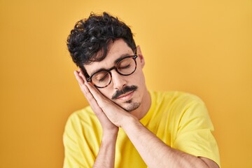 Obraz premium Hispanic man wearing glasses standing over yellow background sleeping tired dreaming and posing with hands together while smiling with closed eyes.