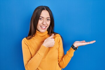 Young brunette woman standing over blue background showing palm hand and doing ok gesture with thumbs up, smiling happy and cheerful