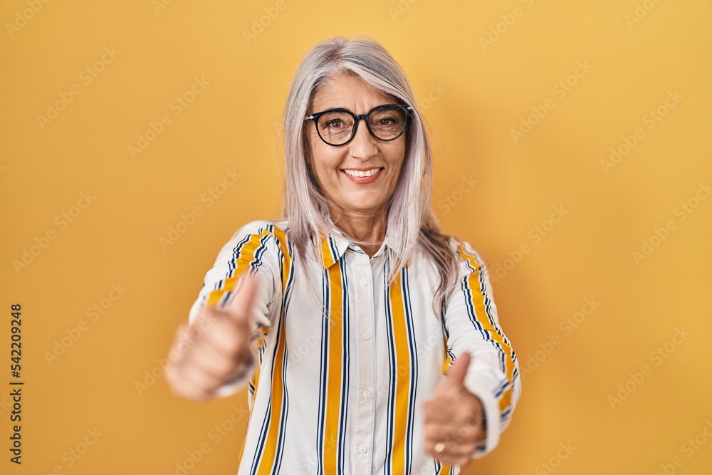 Wall mural Middle age woman with grey hair standing over yellow background wearing glasses approving doing positive gesture with hand, thumbs up smiling and happy for success. winner gesture.