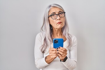 Middle age woman with grey hair using smartphone typing message clueless and confused expression. doubt concept.