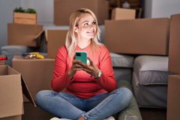 Young blonde woman using smartphone sitting on floor at new home