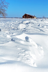 Fototapeta na wymiar Focus on snowdrifts with blurred rural buildings in the background and a blue sky. Winter landscape in Quebec province, Canada