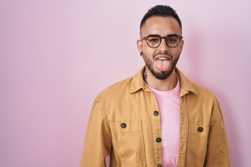 Young hispanic man standing over pink background sticking tongue out happy with funny expression. emotion concept.