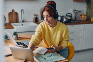Beautiful woman in headphones using technologies while sitting at the kitchen counter