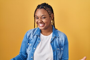 Obraz na płótnie Canvas African american woman with braids standing over yellow background smiling cheerful with open arms as friendly welcome, positive and confident greetings