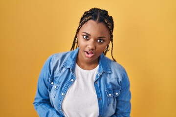 African american woman with braids standing over yellow background in shock face, looking skeptical...