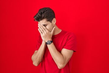 Young hispanic man standing over red background with sad expression covering face with hands while crying. depression concept.