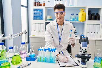 Fototapeta premium Young hispanic man wearing scientist uniform writing on clipboard at laboratory