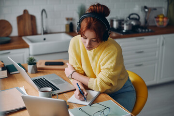 Concentrated woman in headphones making notes in her pad while sitting at the kitchen counter