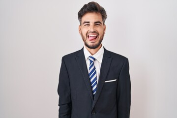 Young hispanic man with tattoos wearing business suit and tie sticking tongue out happy with funny expression. emotion concept.